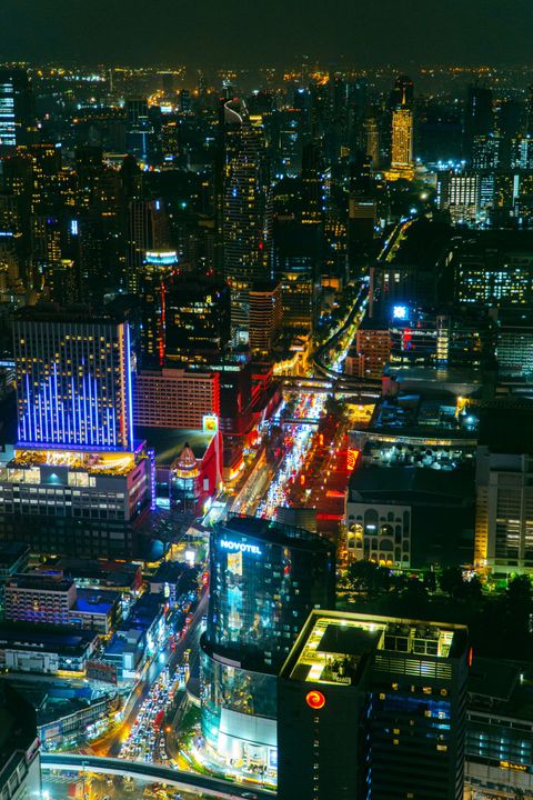 Bangkoks skyline i mörker sedd uppifrån, med Skytrains upplysta spår som skär genom bilden, en Novotel-skylt och bilköer på gatan under