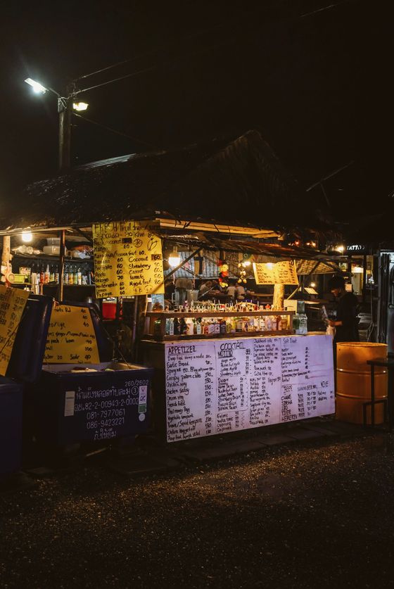 Ett upplyst matstånd på en nattmarknad i Khao Lak, med handskrivna menyer och en rad flaskor på disken