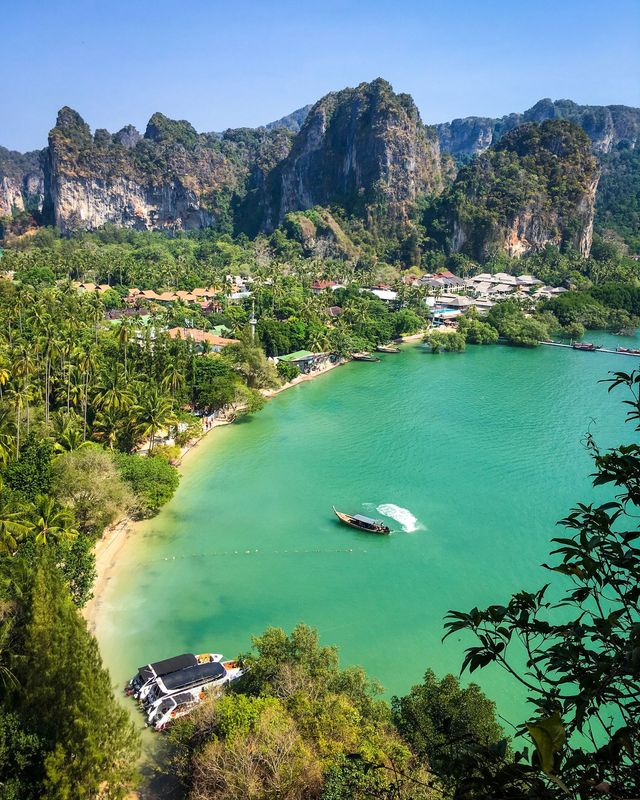Railay-halvön sedd uppifrån, med kalkstensklippor, resorter bland palmerna och en longtailbåt i grönt vatten